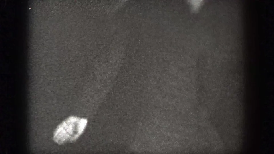 A grainy 8mm frame shows a mostly dark surface with a small bright object, likely part of the same indoor home-movie scene.