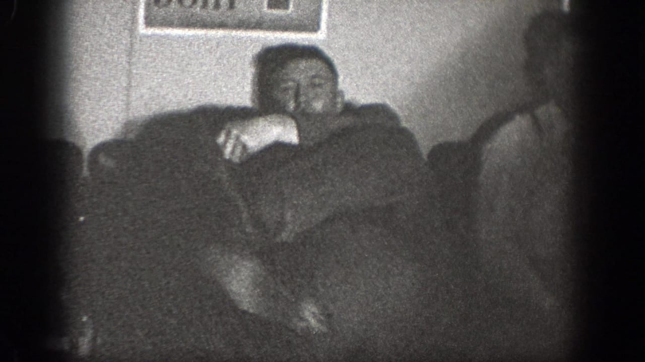 1939 Chelan, Washington home-movie frame showing adult men in a dim interior with upholstered seating and a framed wall picture.