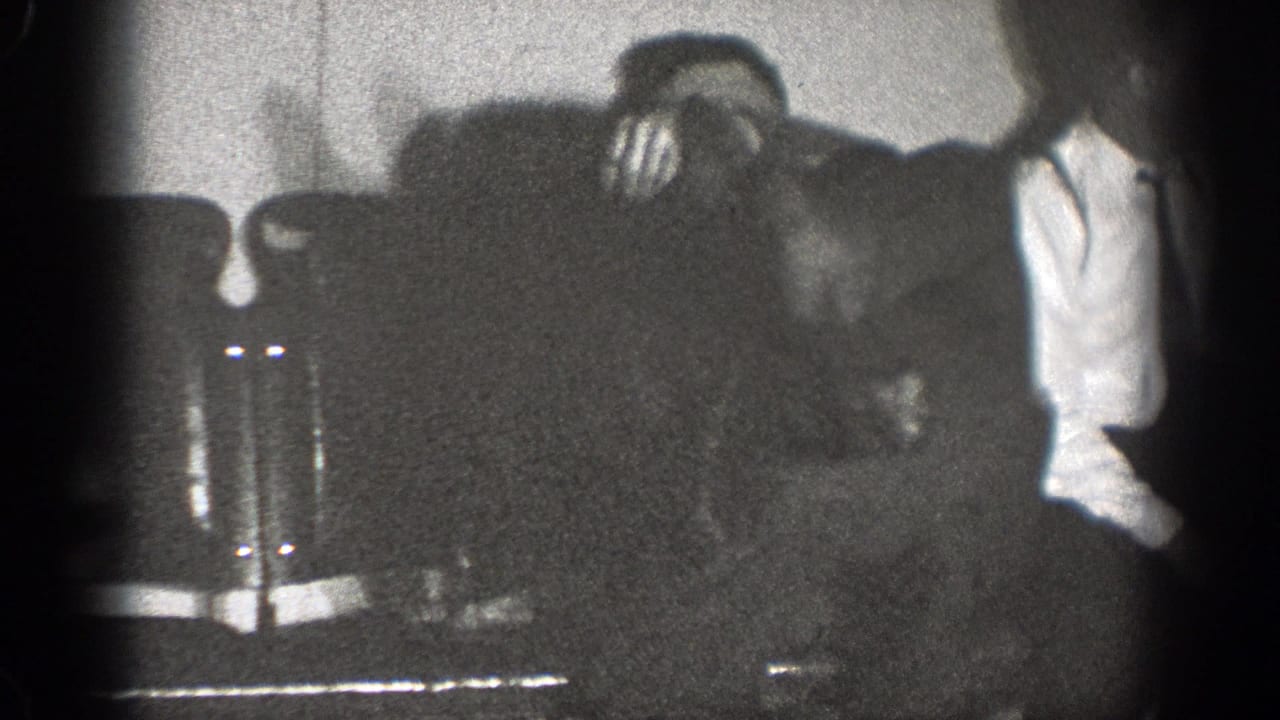 Man in a white shirt stands beside an upholstered chair in a dim room.