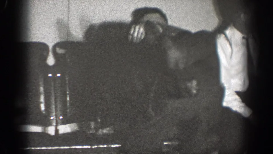 Black-and-white 8mm frame from Chelan showing a standing man in a white shirt near upholstered seating inside a private room.