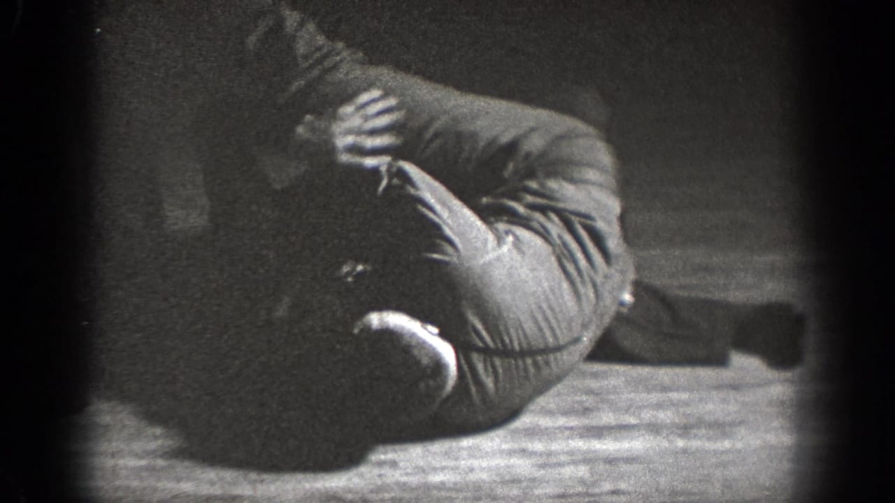Two adult men wrestle on a wooden floor in a dim interior.