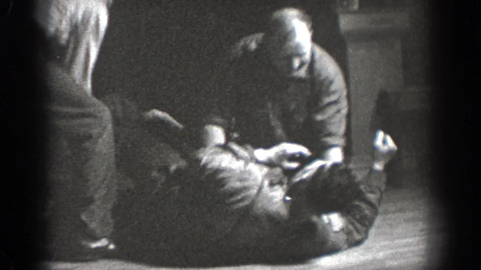 The 1939 home-movie frame shows one man on his back, another crouched over him, and a third person standing at the edge.