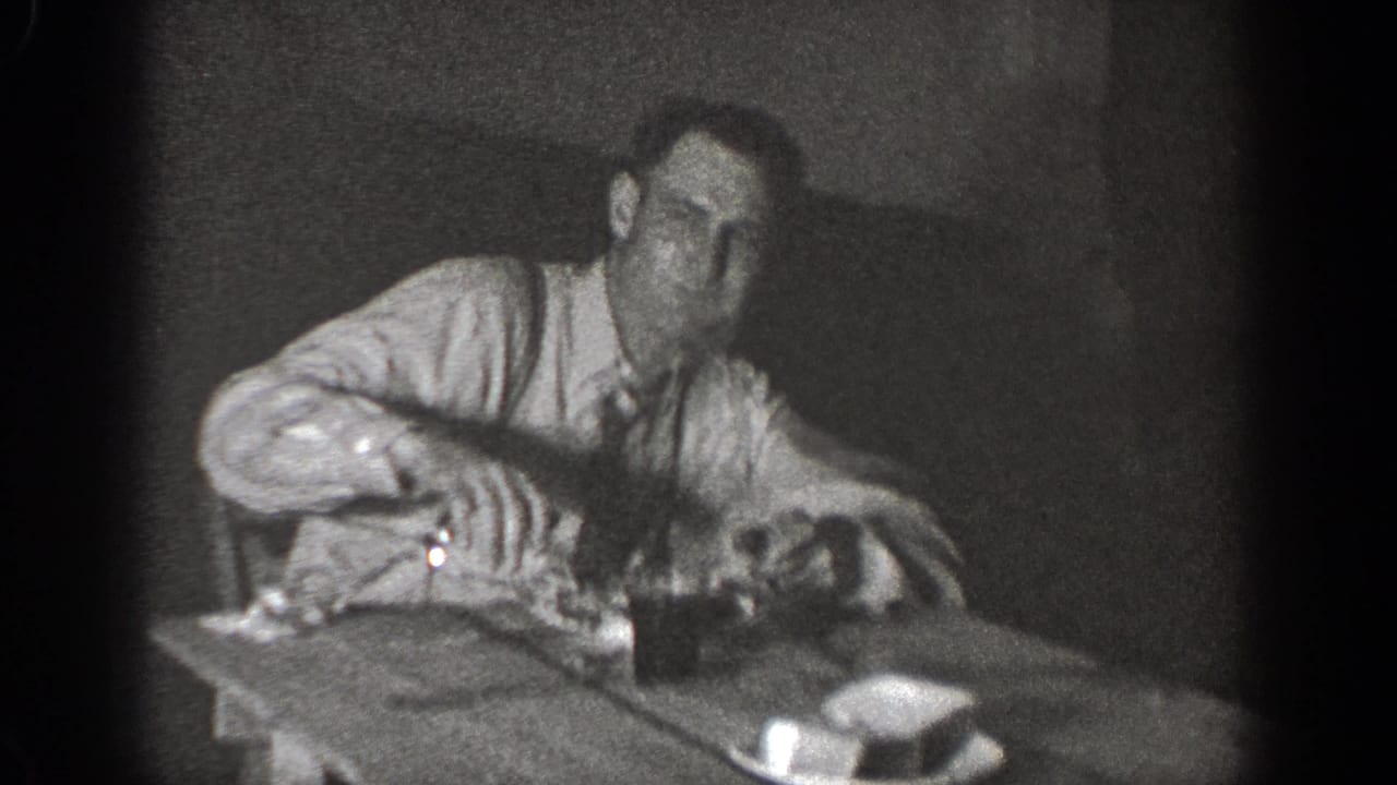 In supported 1939 Chelan footage, a man in a light shirt and tie leans forward at a wooden table with small objects nearby.