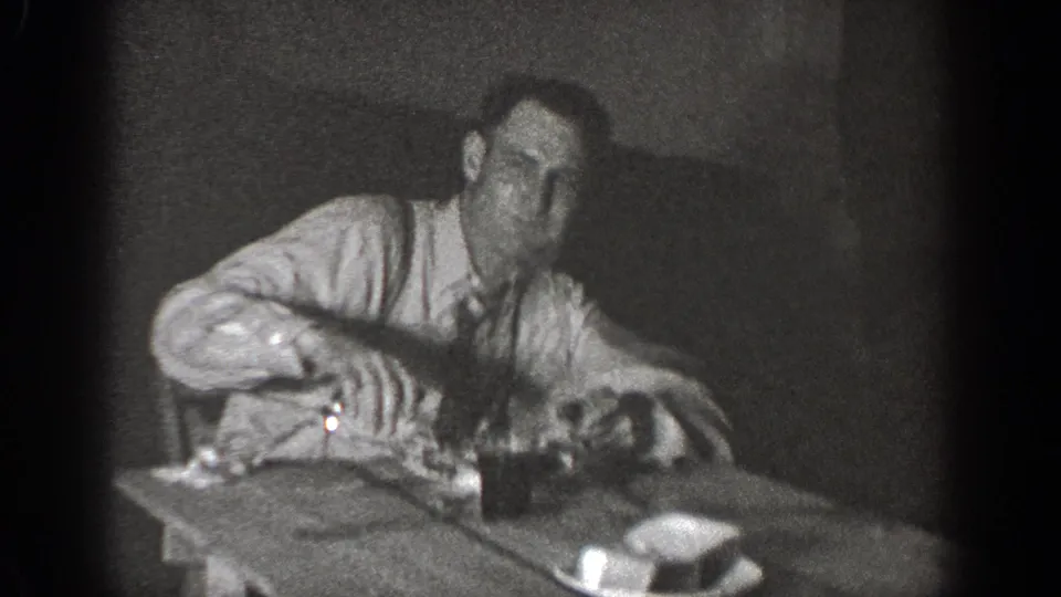 In supported 1939 Chelan footage, a man in a light shirt and tie leans forward at a wooden table with small objects nearby.