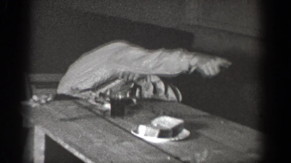 A seated man in a shirt and tie points emphatically across a wooden table in the 1939 Chelan home-movie scene.