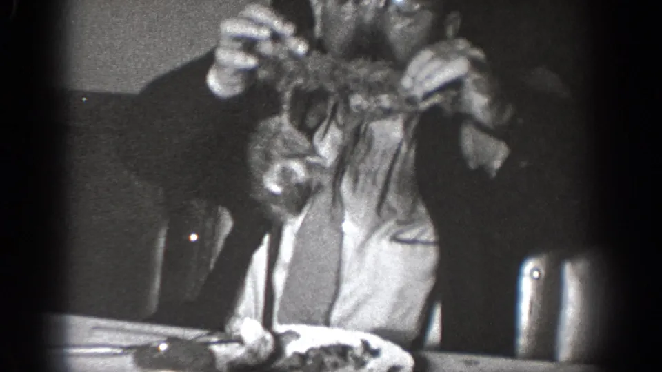 A man with glasses in the Chelan home-movie footage holds a small dark object close to his face while seated at a table.