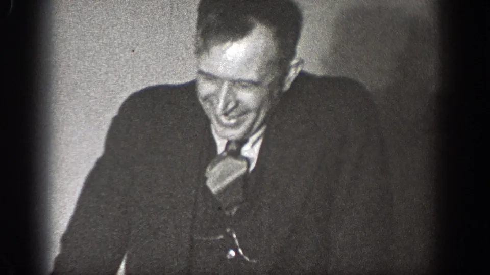 A smiling man in a dark suit and tie sits against a plain interior wall in the supported 1939 Chelan footage.
