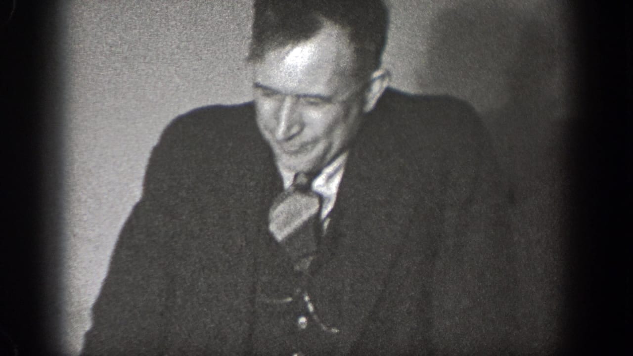 Man in a dark suit and tie laughing in a dim interior.