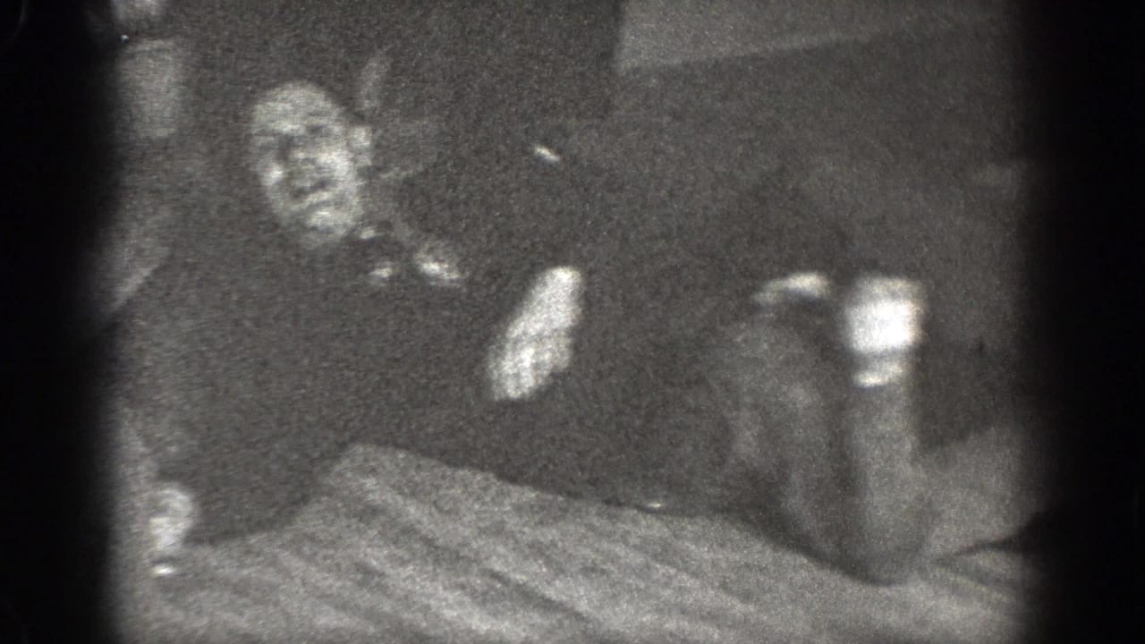 A man appears to recline on a wooden floor near a patterned rug, filmed in grainy black-and-white 8mm in Chelan, Washington.