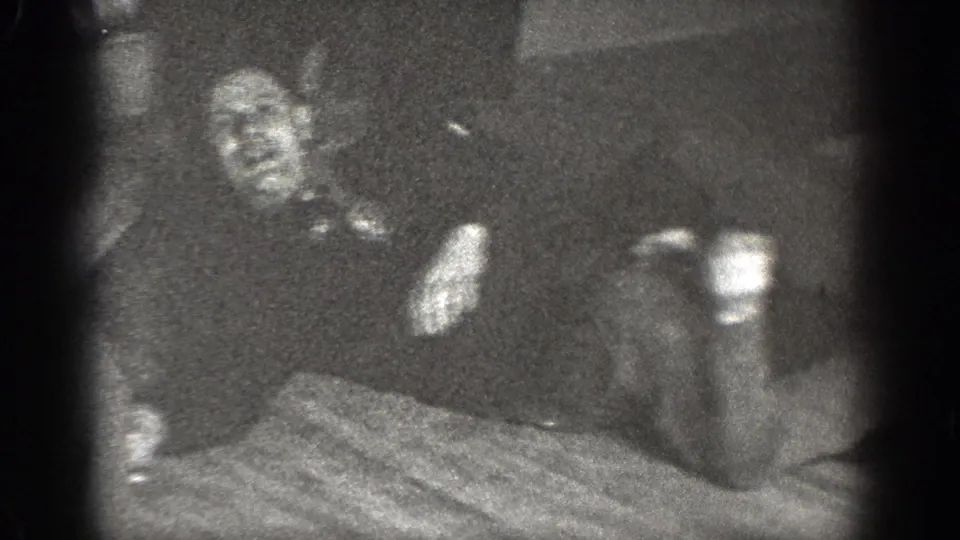 A man appears to recline on a wooden floor near a patterned rug, filmed in grainy black-and-white 8mm in Chelan, Washington.
