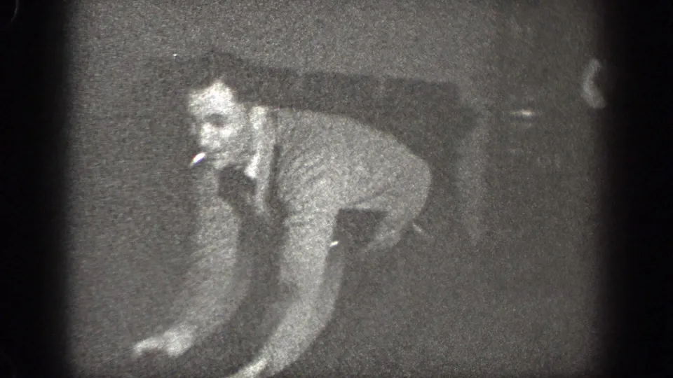 An adult man in light clothing is down on hands and knees on a wooden floor, with another person partly visible at the edge of the frame.
