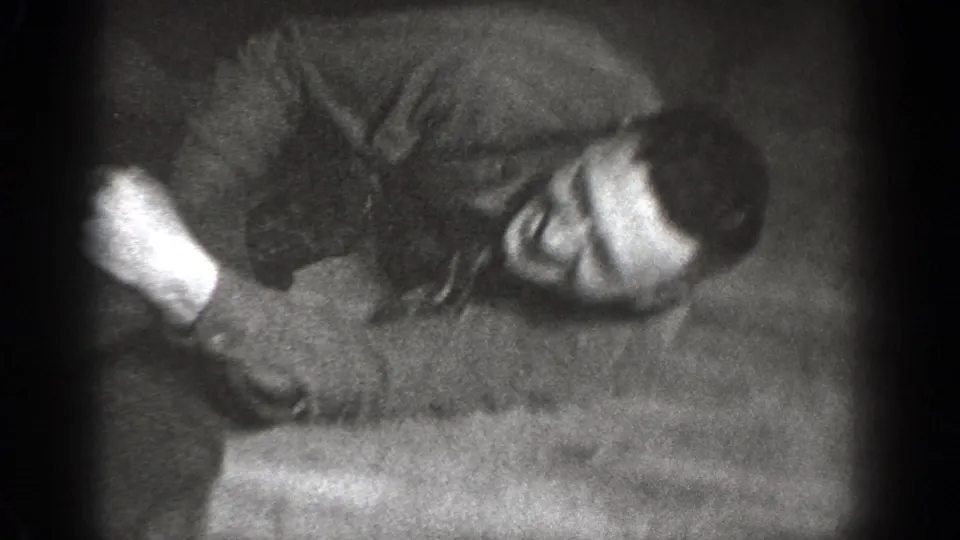 A suited man lies sideways on the floor, smiling or laughing as the home-movie camera catches the playful moment.