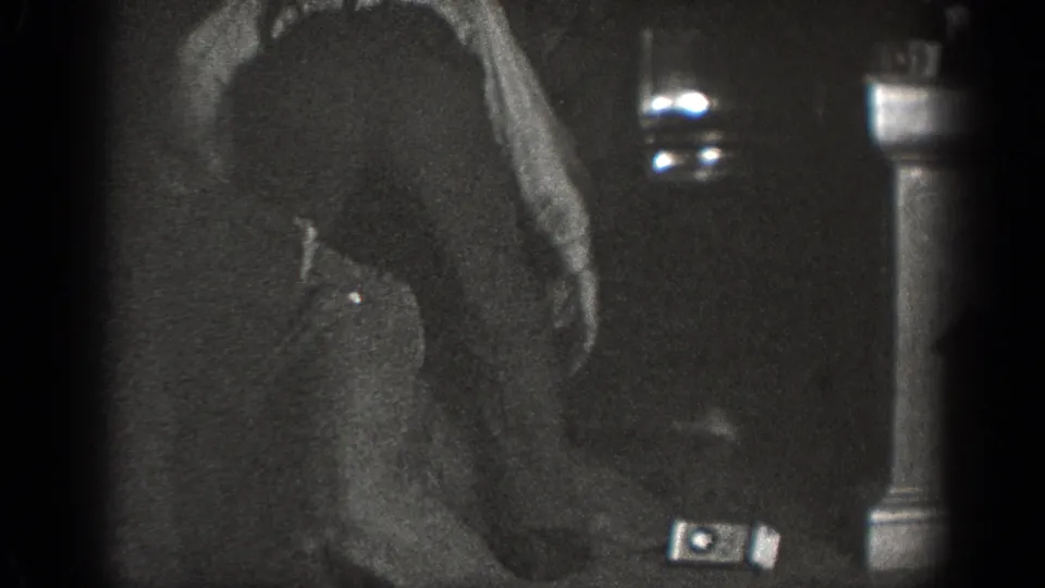 A dark-clothed adult bends forward in the dim 1939 Chelan interior, with a small bright object visible near the right edge.