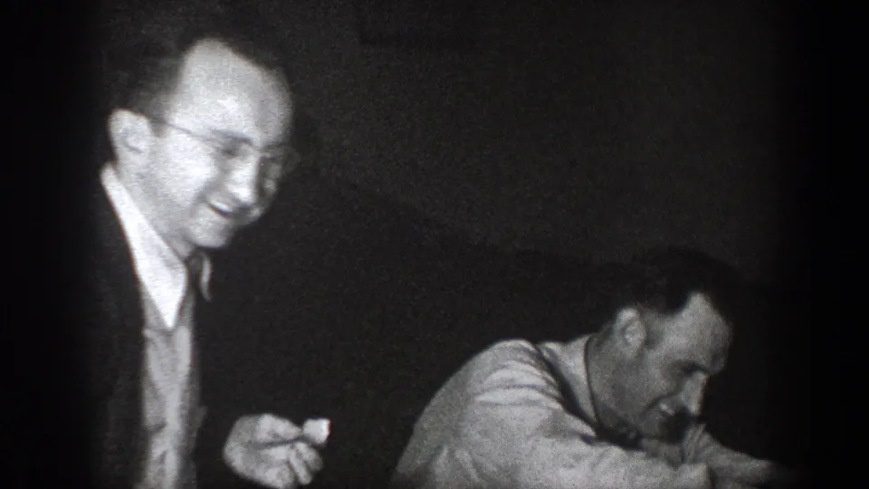 A smiling man with glasses stands beside another man leaning forward in the 1939 Chelan indoor gathering.