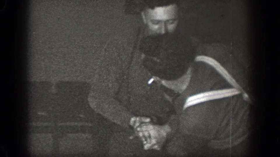 A bearded man stands over another man holding a small can or cup in the dim 1939 Chelan home-movie scene.