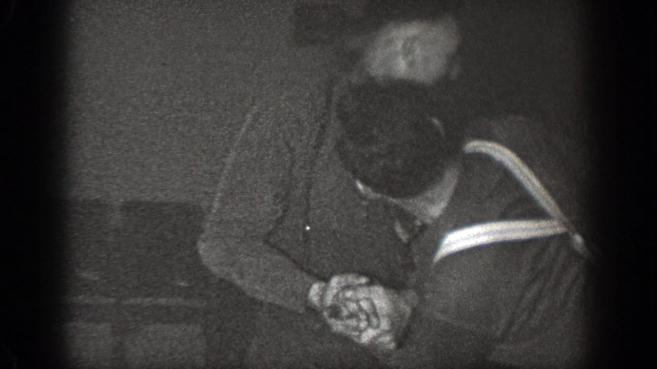 Two adult men in dark clothing lean together with hands clasped in a dim room.
