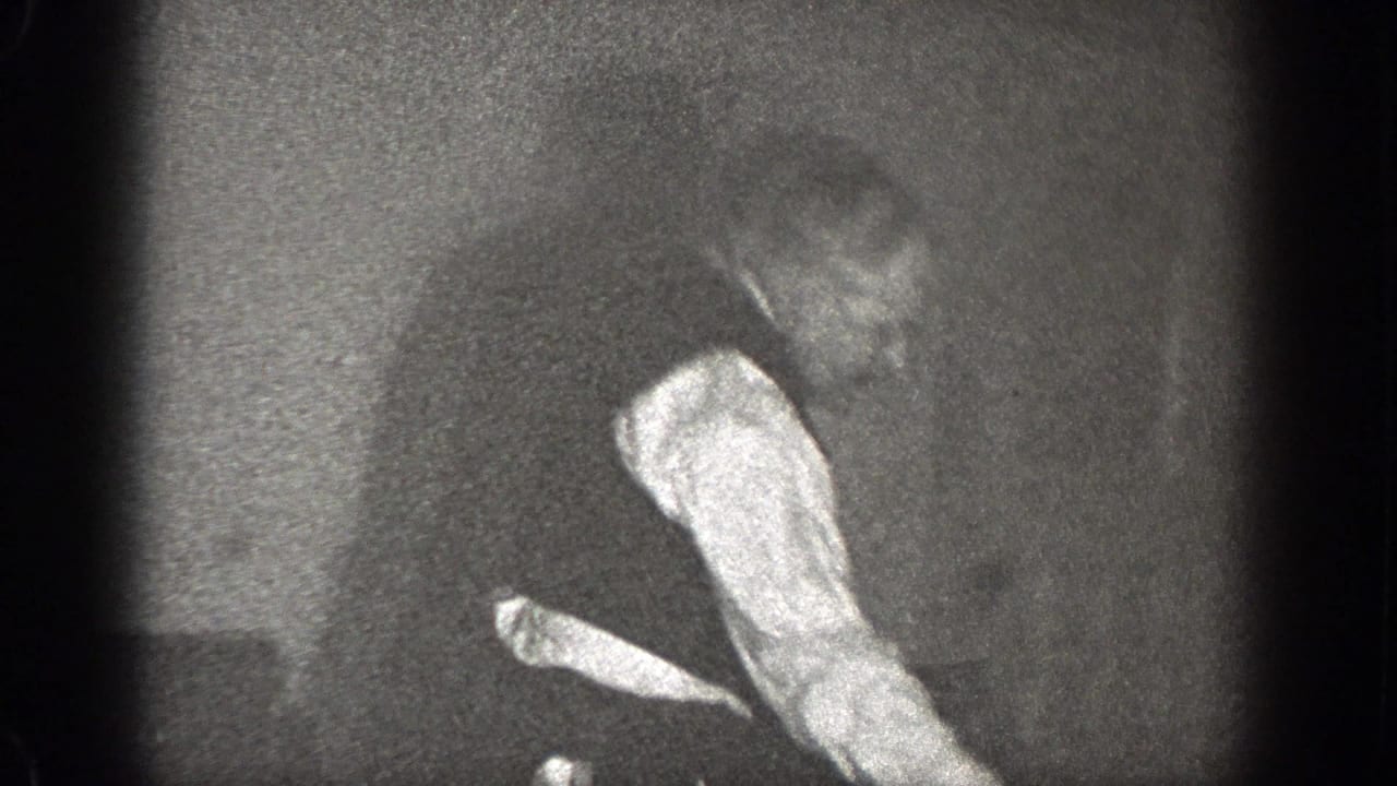 Man in shirtsleeves and vest bends or walks near a dim wall.