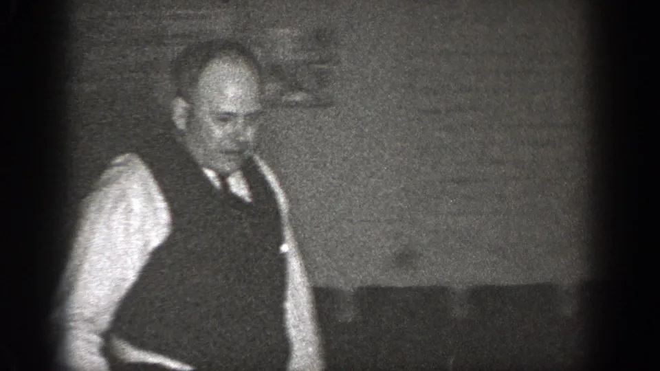 A balding man in vest and tie stands in the room, with a partially visible marked box or sign behind him.