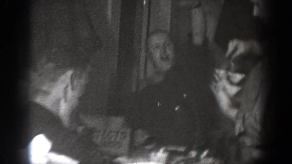 Adults gather around a table in the dim Chelan interior, with faces and shirtsleeves visible in soft black-and-white footage.