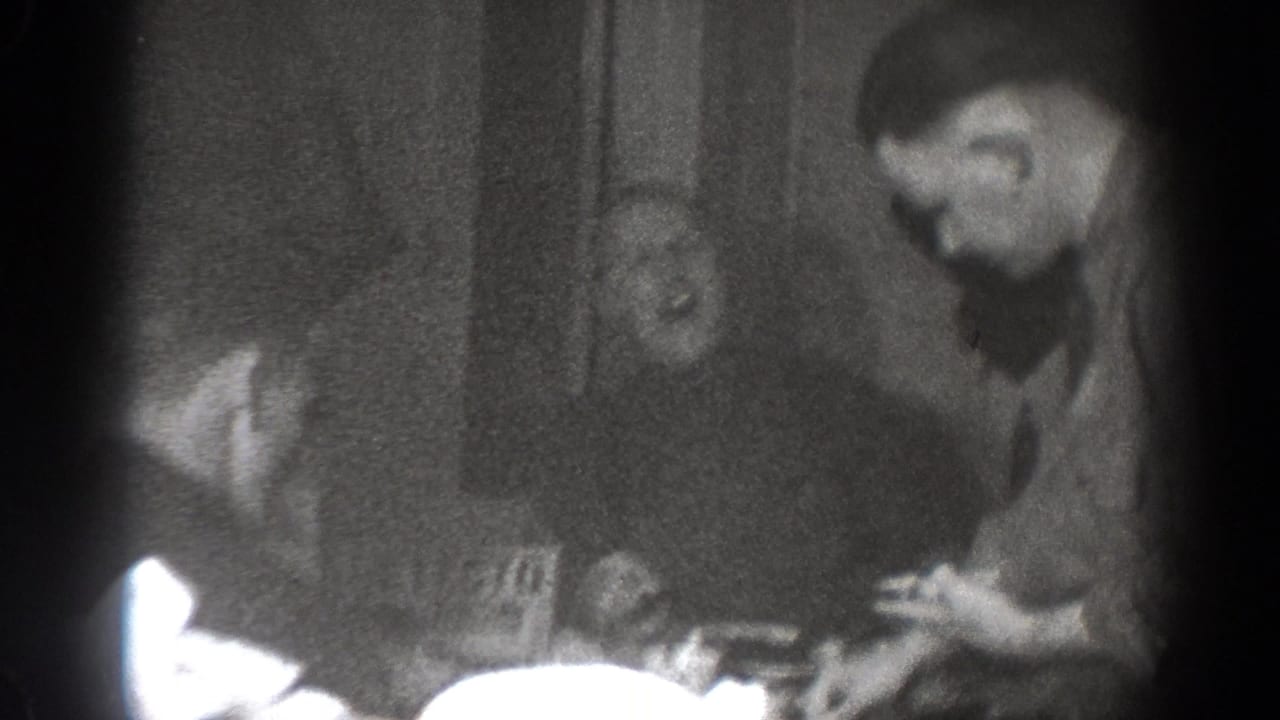 Men sit around a table in a dim room, with one man leaning forward near plates and cups.