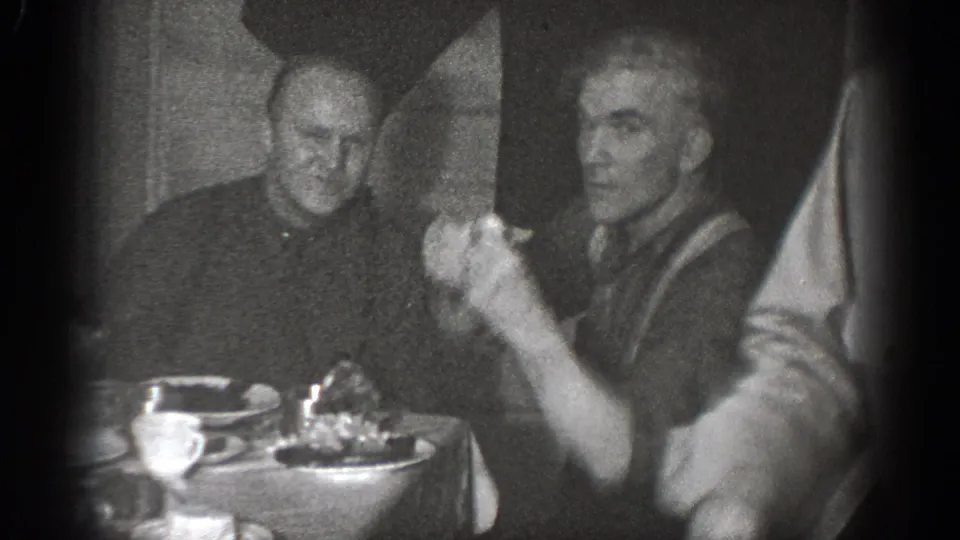 Adult men sit beside plates and cups in a 1939 Chelan home-movie frame, with one man looking toward the camera.