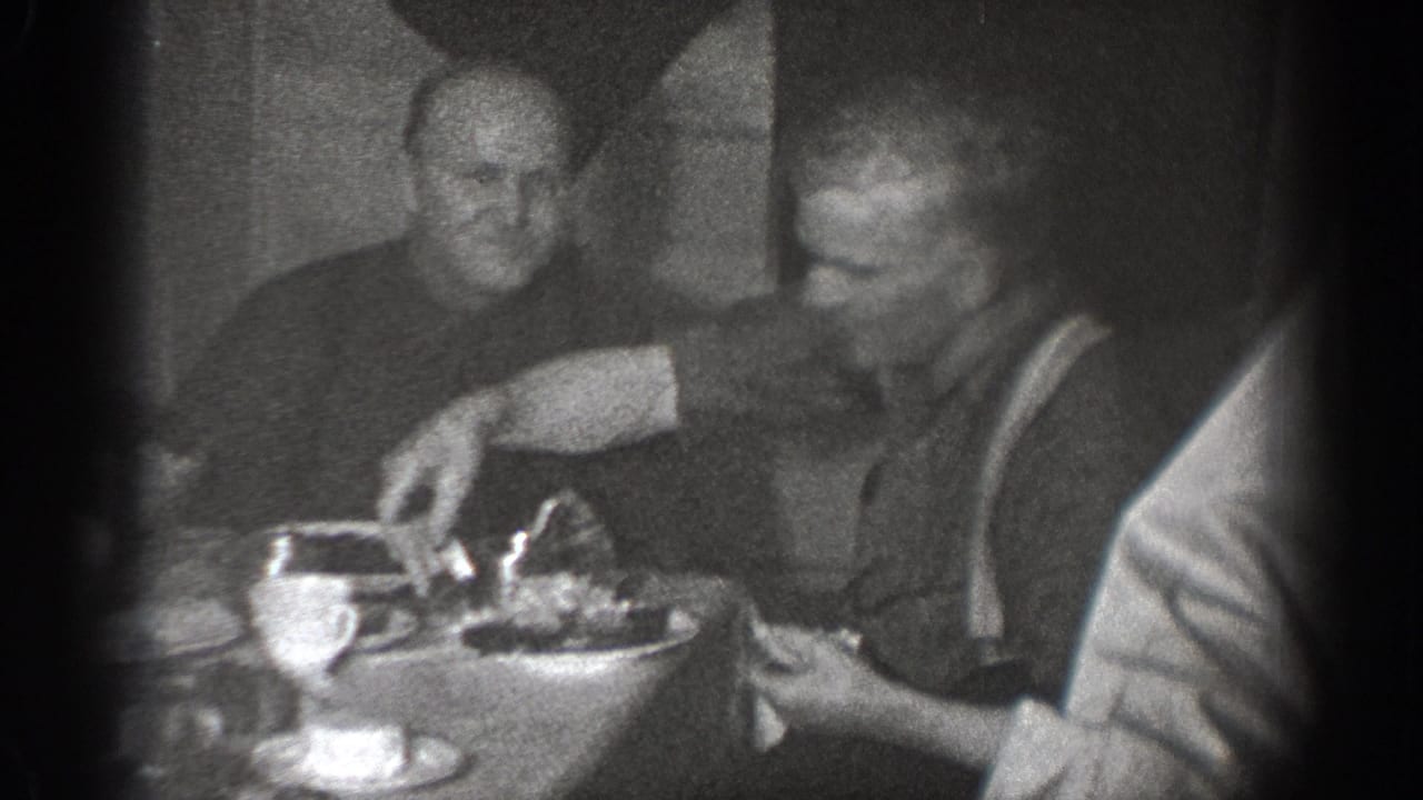 In Chelan, Washington home-movie footage from 1939, two men sit at a dining table with plates and cups in a dark room.