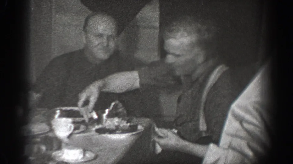 In Chelan, Washington home-movie footage from 1939, two men sit at a dining table with plates and cups in a dark room.