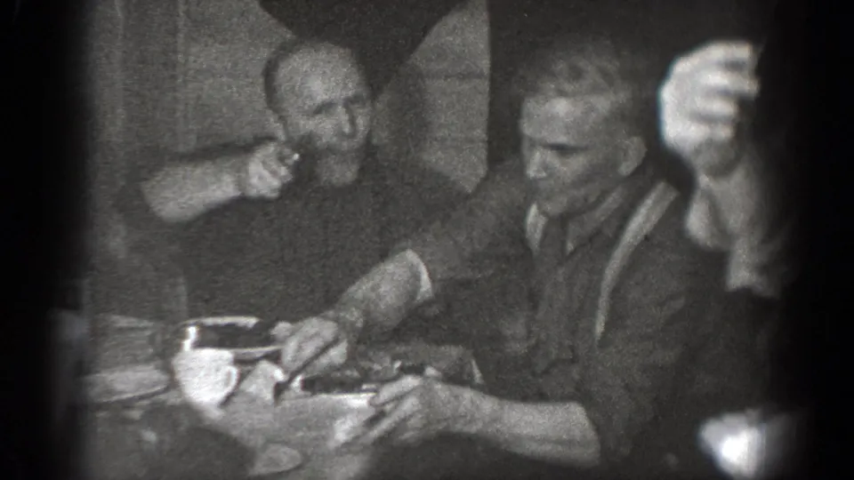 In the 1939 Chelan home movie, one seated man points at the camera as another man handles food at the table.