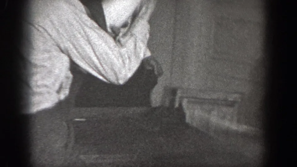 1939 Chelan home-movie frame showing a cropped adult man in a light shirt and suspenders inside a dim private room.