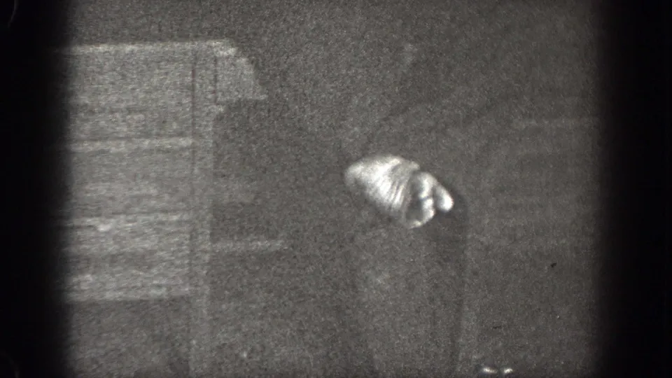 In a grainy 1939 Chelan interior, a shirted man bends near a wall or cabinet while handling a sock or foot.