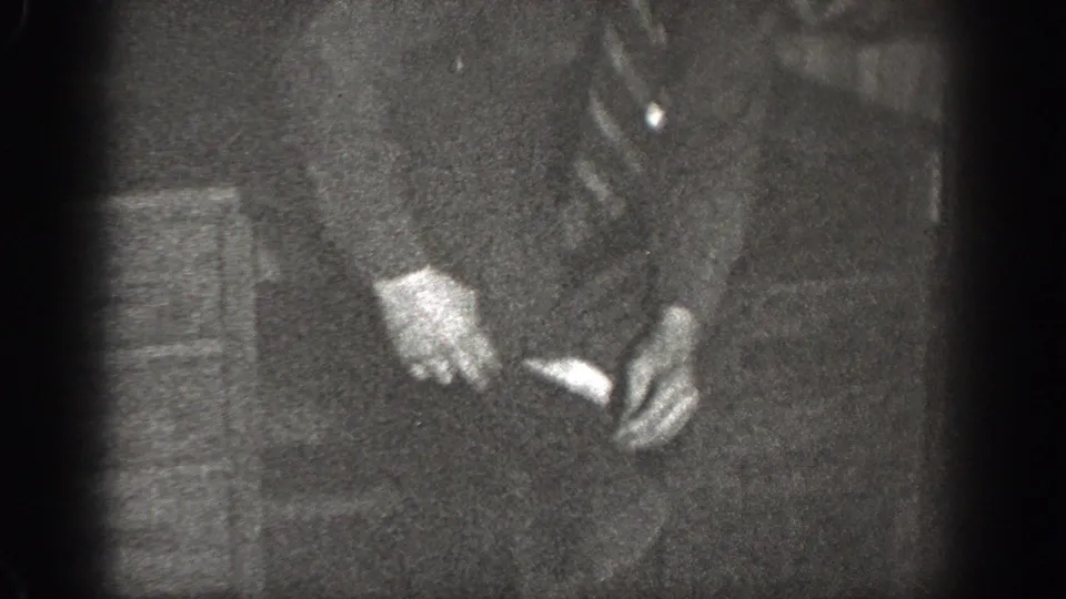 The seated man in a dark suit appears to handle a small light object while seated indoors in the Chelan home-movie footage.