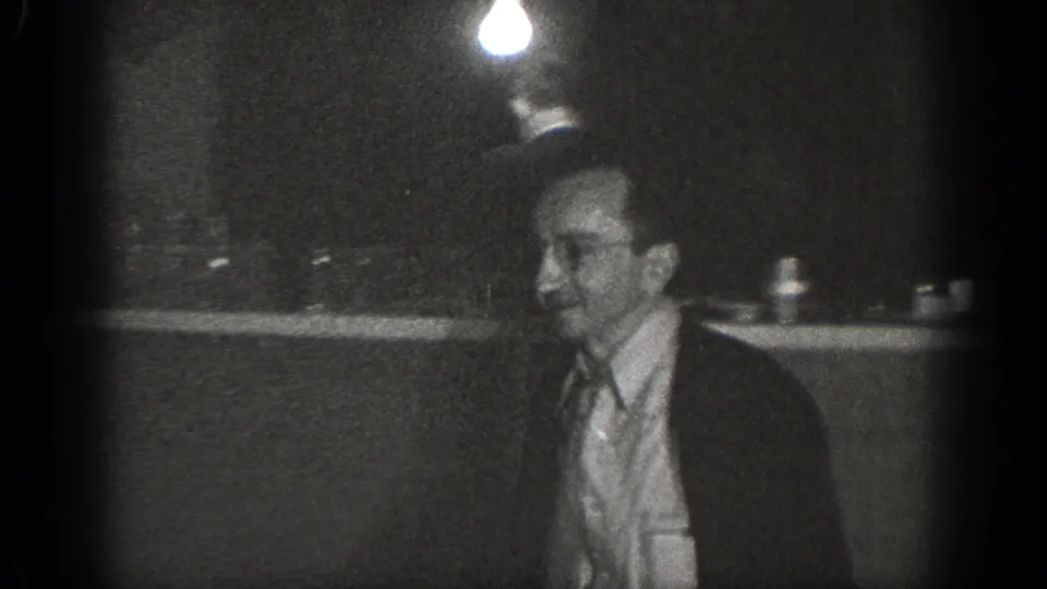 A second adult man in a light shirt briefly appears in the dim Chelan interior, seen in profile with soft focus and visible grain.