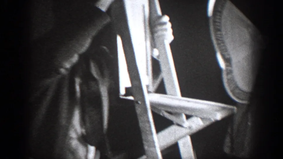 A partial close view shows a person beside a wooden chair, with a dark coat and cropped body details in the grainy 1939 interior footage.