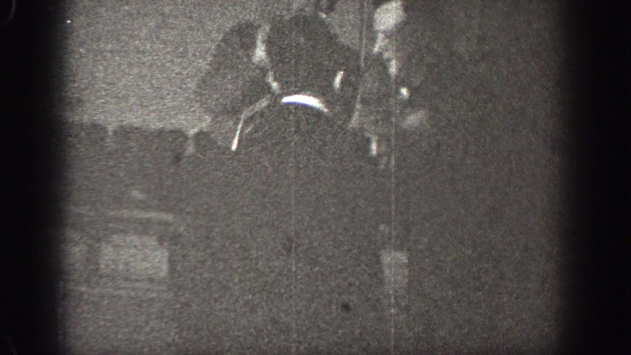 In Chelan, Washington 1939 home-movie footage, two adult men appear close together in a dim interior with heavy grain and soft focus.