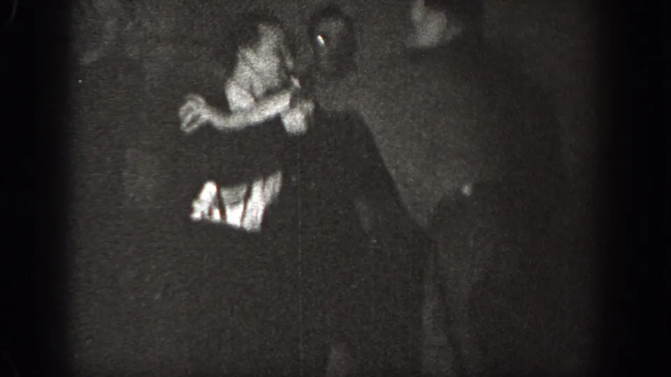 A 1939 Chelan home-movie frame shows one adult man leaning forward while another in a dark suit remains close in a dim indoor setting.