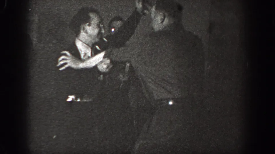 In a grainy 1939 Chelan interior frame, two adult men face each other closely, with one holding a cigarette during an ambiguous tense exchange.