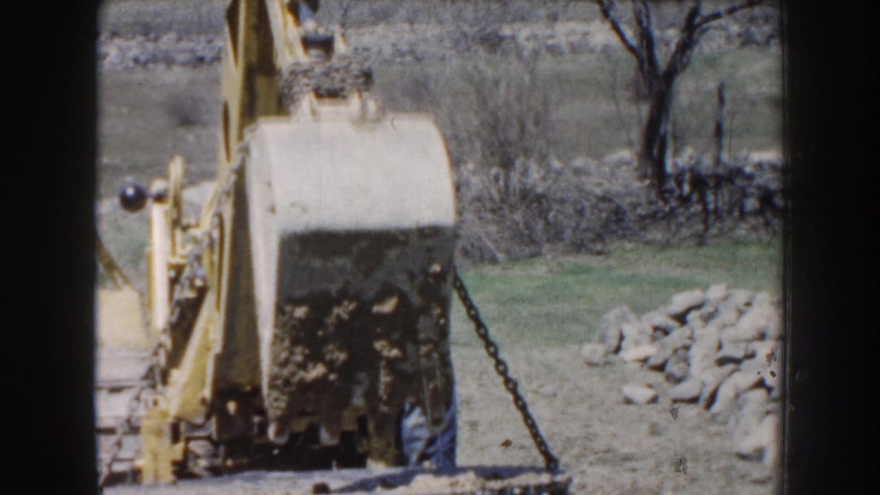 1959 Nebraska 8mm frame showing a yellow tractor or loader raising a concrete block with chain at a rural outdoor work site.