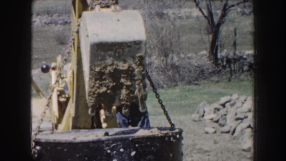 1959 Nebraska home-movie frame with a chain-suspended concrete block held by a yellow tractor near a dark bucket or form.