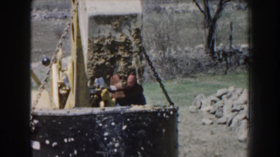 1959 Nebraska 8mm frame showing a worker guiding equipment near a tractor and chain-lifted concrete block outdoors.