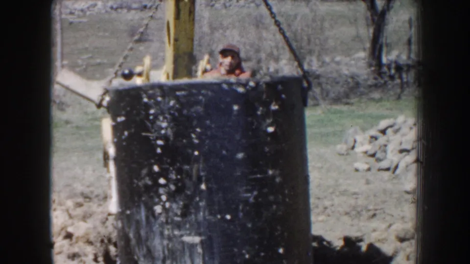 1959 Nebraska home-movie frame with a man in work clothes visible behind a black bucket or form near tractor chain rigging.