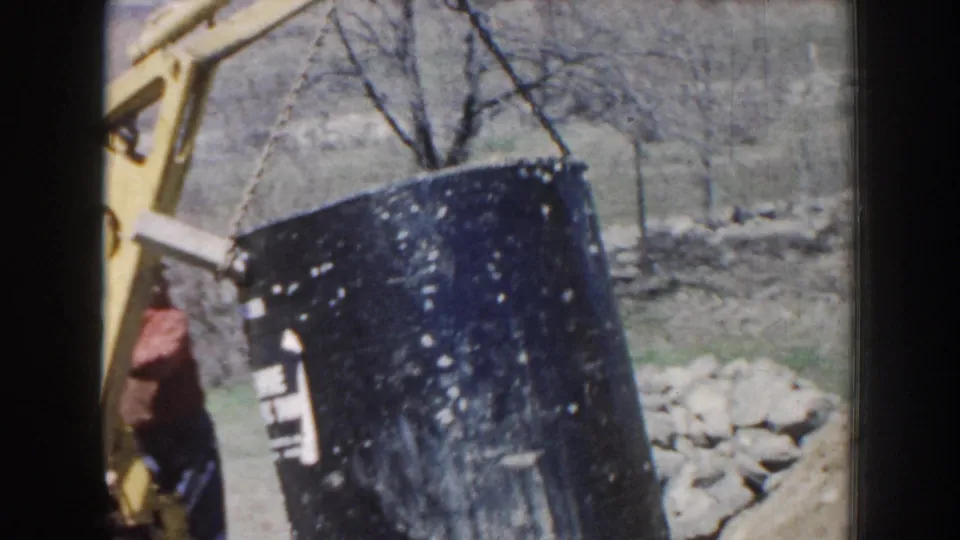 A worker in a red shirt is partly visible as the yellow crane tilts the black septic tank over rural Nebraska ground.