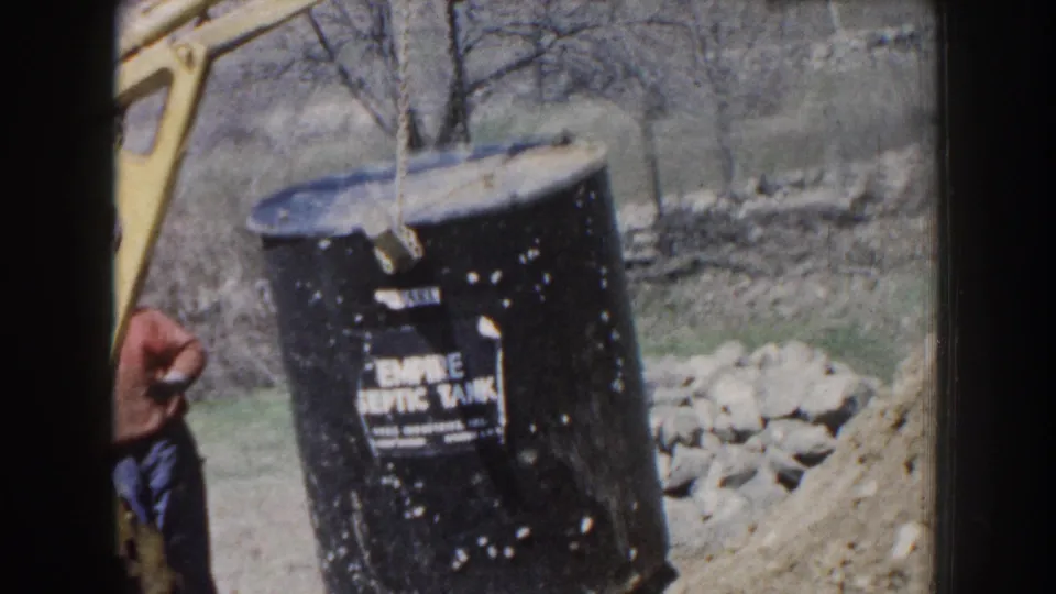 The black tank with an Empire Septic Tank label is lifted by a yellow crane above a dug trench in rural Nebraska footage.