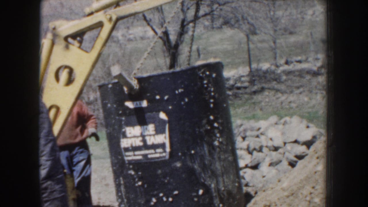 Nebraska 1959 home-movie frame showing an adult worker near a black septic tank being lifted by yellow equipment at a wooded work site.