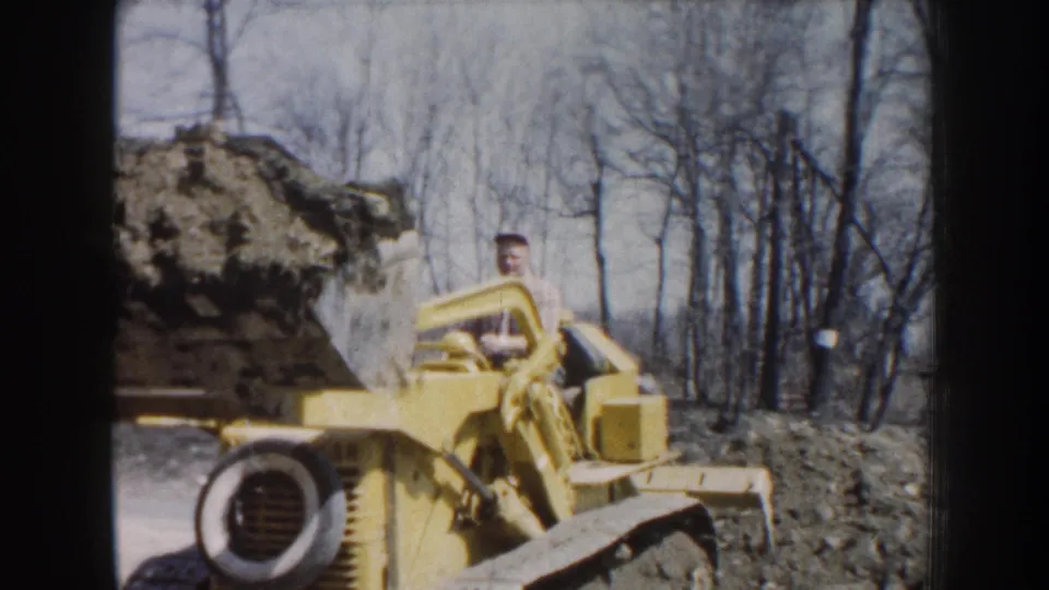 Yellow construction equipment operates in a wooded Nebraska work area, with an adult operator visible and a large load raised above the ground.