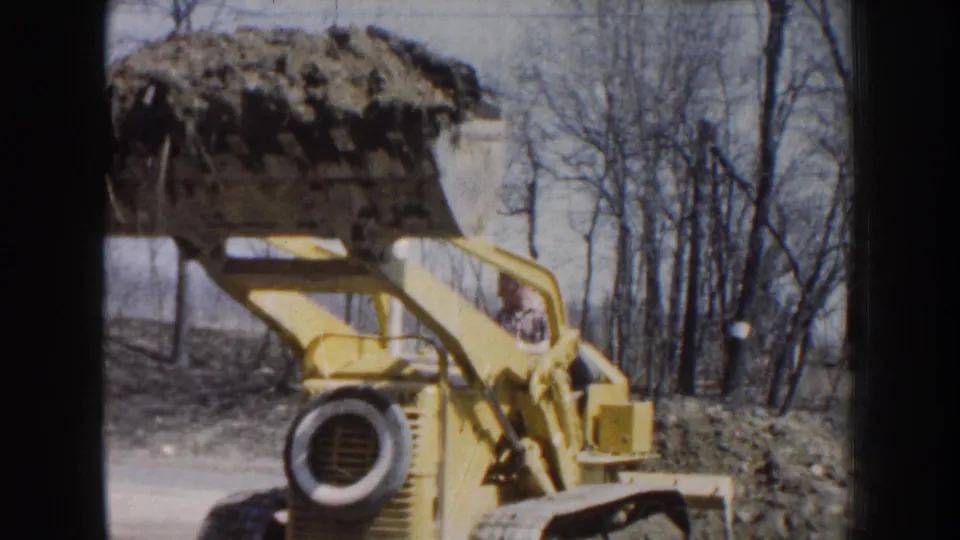 The yellow machine holds its bucket raised over a rural Nebraska work site, with bare trees and disturbed soil in the background.