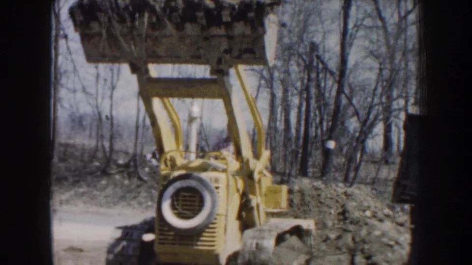 A yellow loader-crane faces the camera with its bucket lifted high at a wooded Nebraska construction site in 1959 home-movie footage.