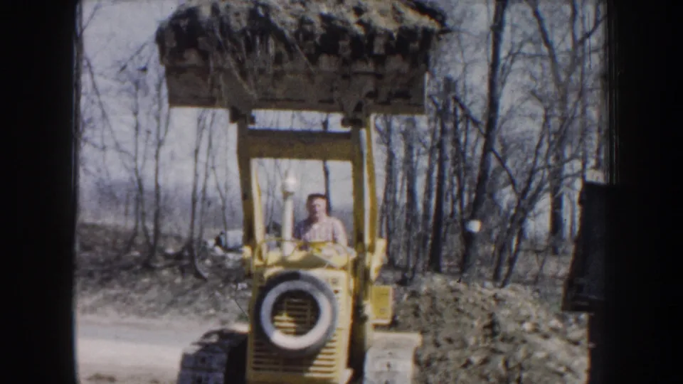 Nebraska 1959 home-movie frame showing a man operating a yellow front-end loader with a raised bucket in a leafless wooded area.