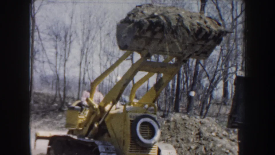 Nebraska 1959 frame with a yellow loader seen from the side, its bucket raised and a worker partly visible at the controls.