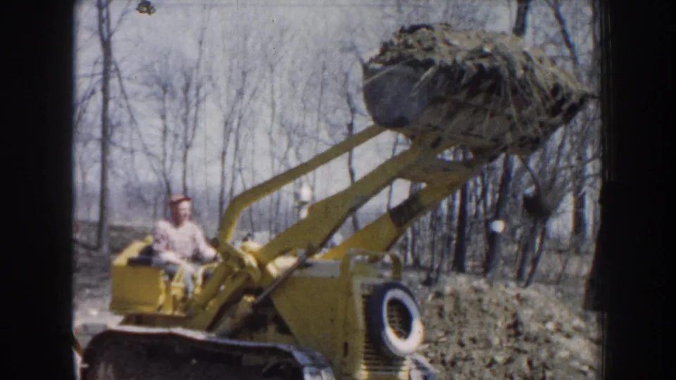 Nebraska 1959 home-movie frame showing the operator in a cap seated on a yellow loader beneath a raised debris-filled bucket.