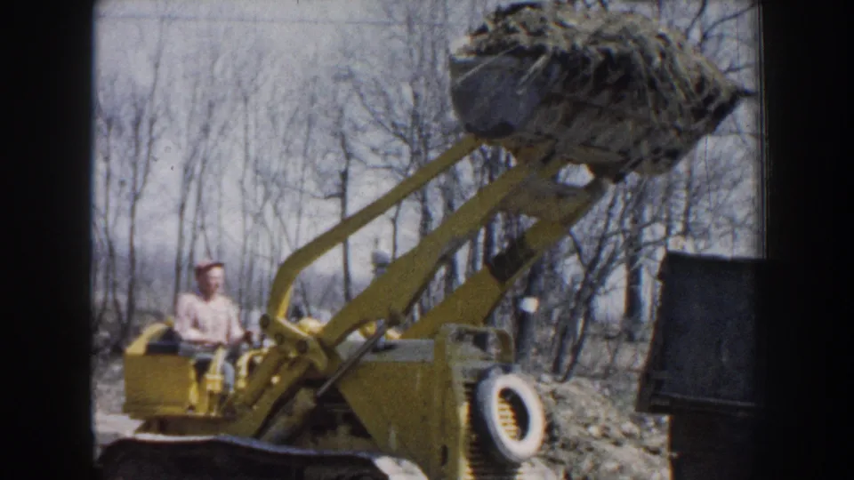 Nebraska 1959 frame of a yellow front-end loader in profile with a seated worker and raised bucket against bare trees.